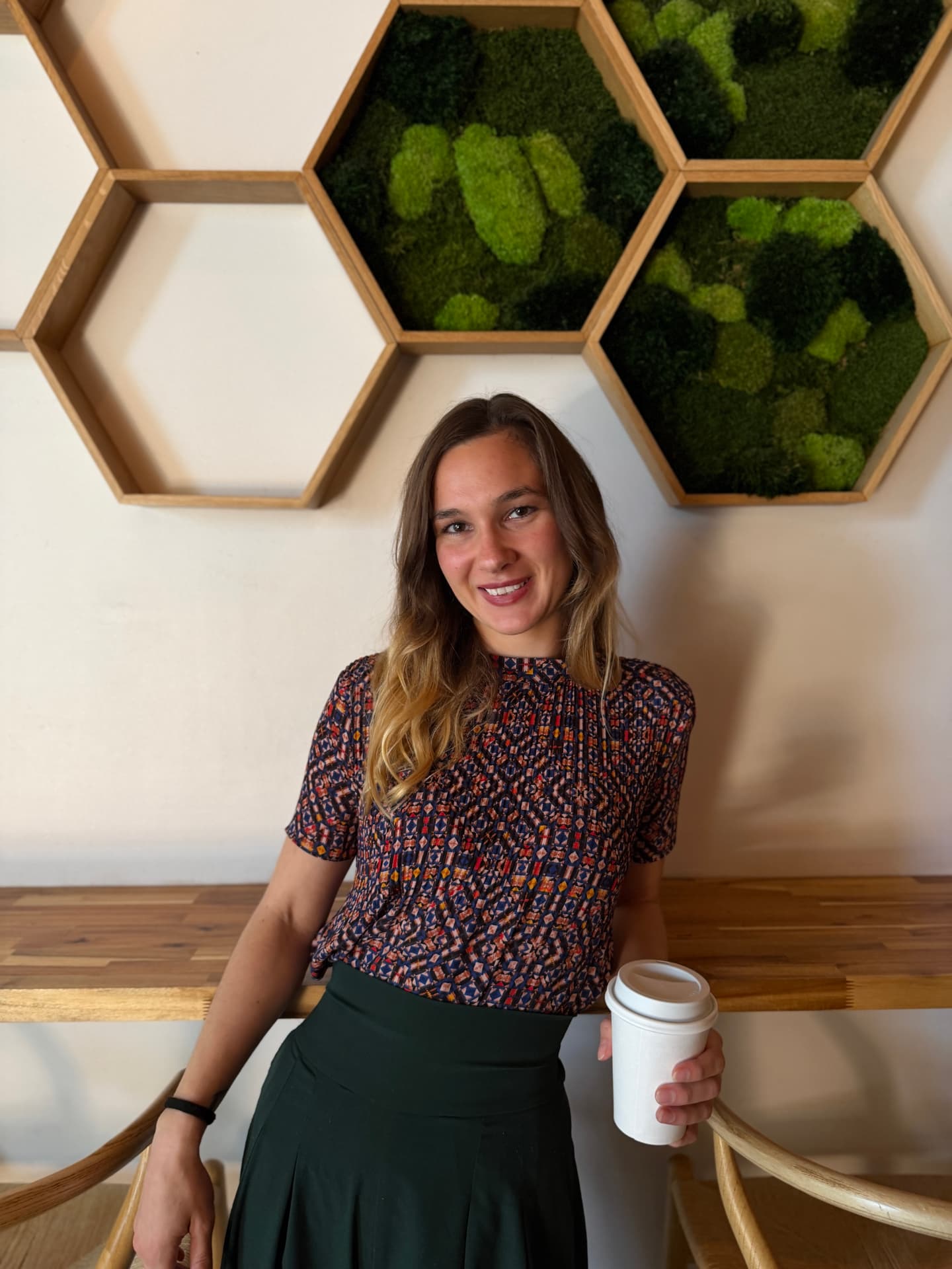 Haile smiling, holding a coffee cup in front of a moss wall